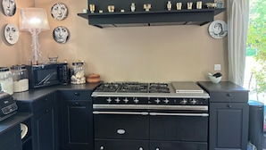 Een keuken met een fornuis, magnetron en kastjes. Er hangen decoratieve borden aan de muur en een raam met uitzicht op groen.