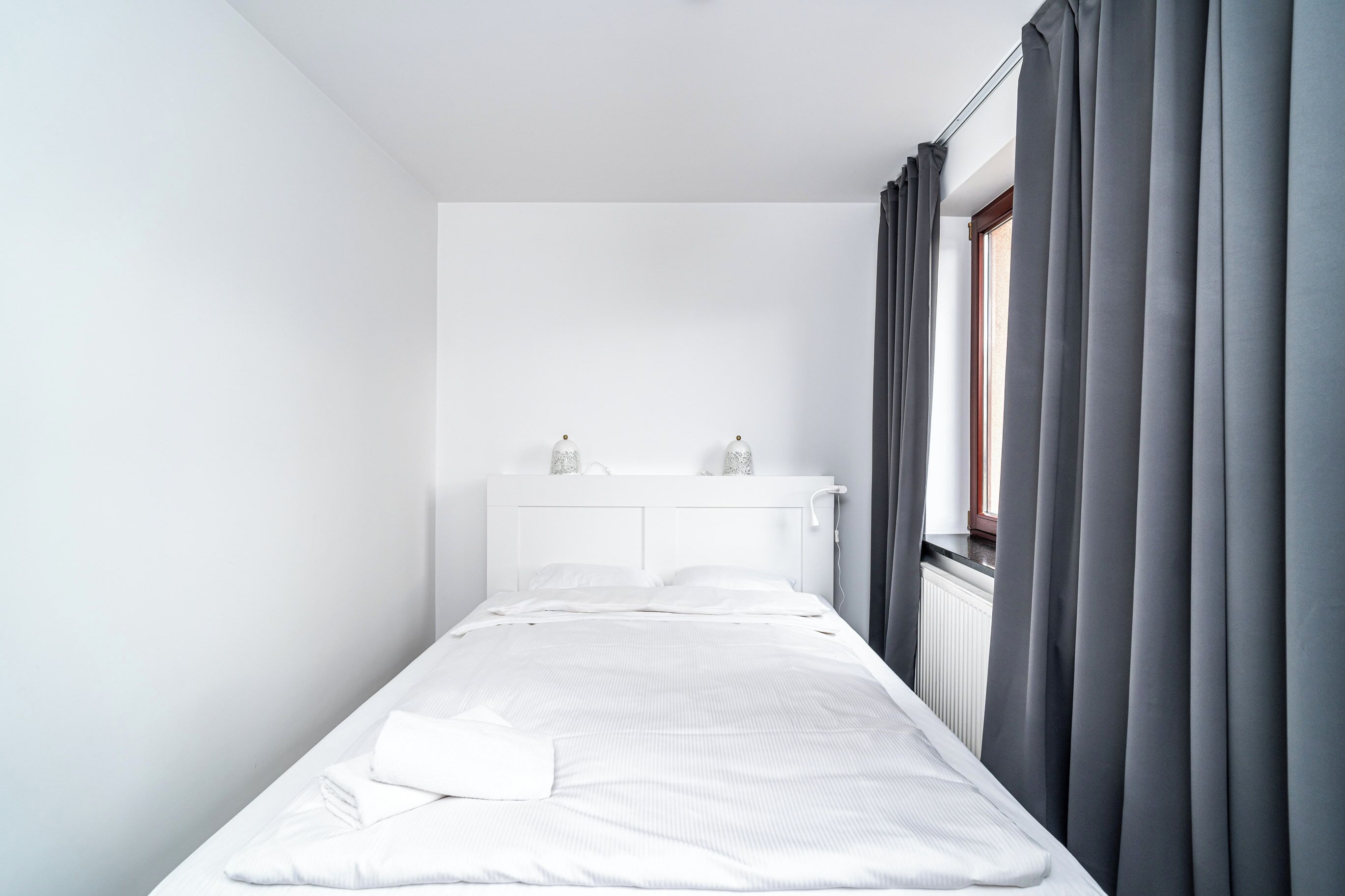 A small, neatly made bed in a minimalistic room with a white headboard, a window with curtains, and a radiator.