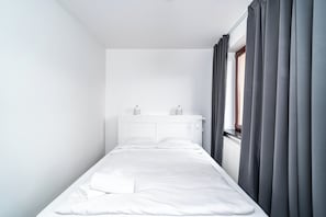 A small, neatly made bed in a minimalistic room with a white headboard, a window with curtains, and a radiator. - RentPlanet - Apartament Zamoyskiego (Kraków)