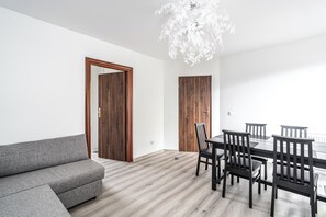 Uma sala de estar moderna com um sofá cinzento, mesa de jantar de madeira com cadeiras e um candeeiro de teto branco.