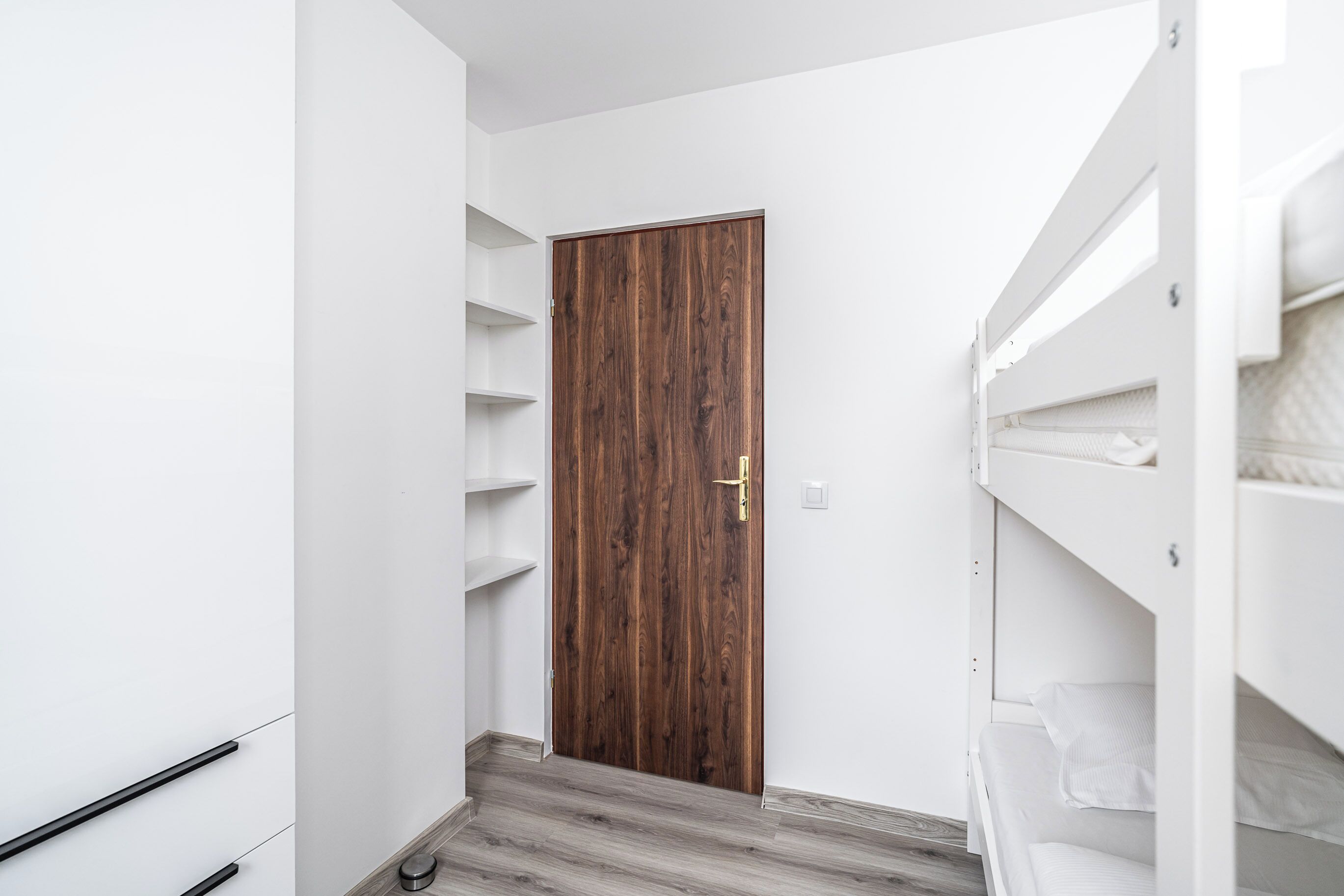 Un corridoio stretto con una porta in legno, pareti bianche e un letto a castello con biancheria bianca.