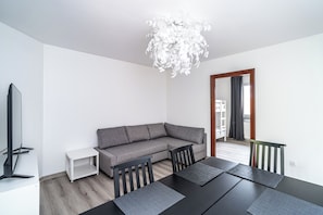 A modern living room with a grey sofa, a dining table with chairs, a flat-screen TV, a white side table, and a chandelier. - RentPlanet - Apartament Zamoyskiego (Kraków)