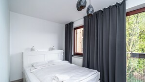 Un dormitorio con cama, ventana con cortinas, radiador y vista a la vegetación exterior.