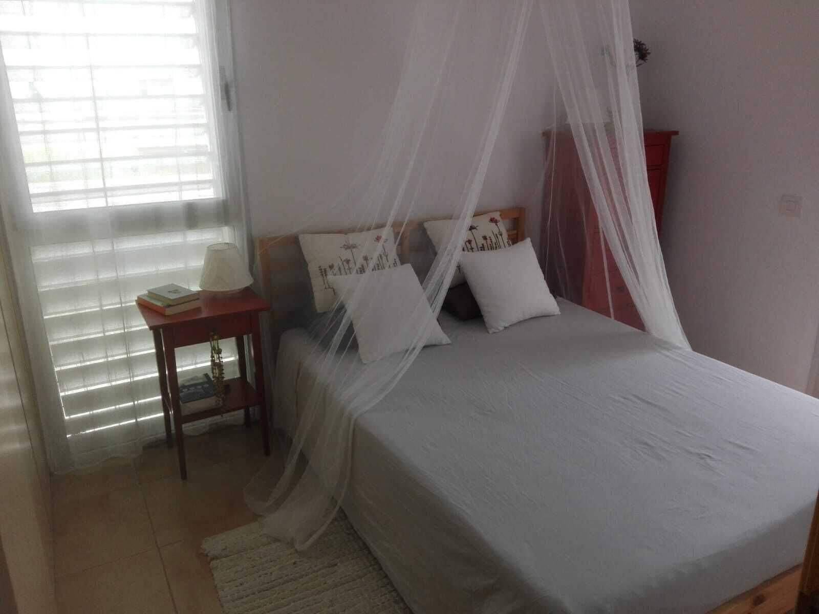 Een slaapkamer met een bed, nachtkastje, lamp en raam met jaloezieën.