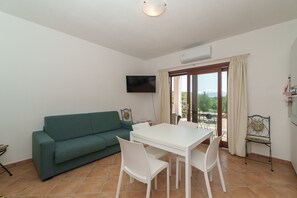Ein Wohnzimmer mit einem grünen Sofa, einem weißen Esstisch mit Stühlen, einem an der Wand befestigten Fernseher und einem Balkon mit Blick.