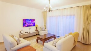 Una sala de estar con un juego de sofá, una mesa de centro, un televisor montado en la pared, una araña y un amplio ventanal con cortinas.