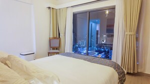 Habitación de hotel con una cama grande, una silla, una mesita y vistas a un paisaje urbano nocturno.