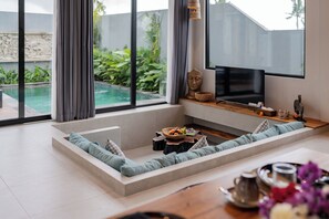 Ein modernes Wohnzimmer mit großem Fenster, einem Flachbildfernseher, einer integrierten Sitzecke und einem kleinen Pool, der durch das Fenster sichtbar ist.