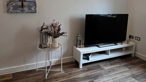 Una sala de estar moderna con un televisor de pantalla plana sobre un mueble blanco, un jarrón decorativo con flores y una lámpara de pared.