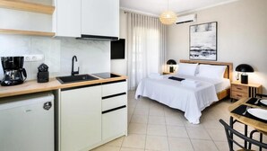 Habitación de hotel pequeña con kitchenette, cama con ropa de cama blanca, mesita de noche con lámpara y un cuadro en la pared.