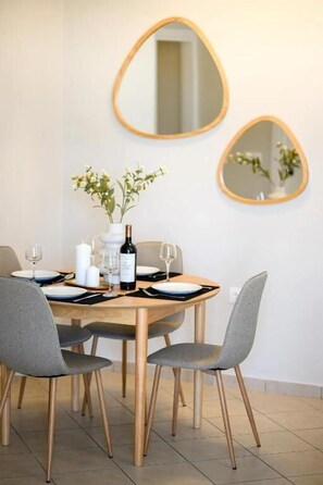 Un comedor con una mesa redonda, cuatro sillas grises, dos espejos colgados en la pared, un jarrón con flores, una botella de vino y una vela.