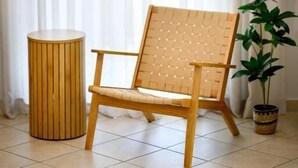 Una habitación con una silla de madera, un taburete redondo de madera, una planta en maceta y un gran ventanal con cortinas translúcidas.