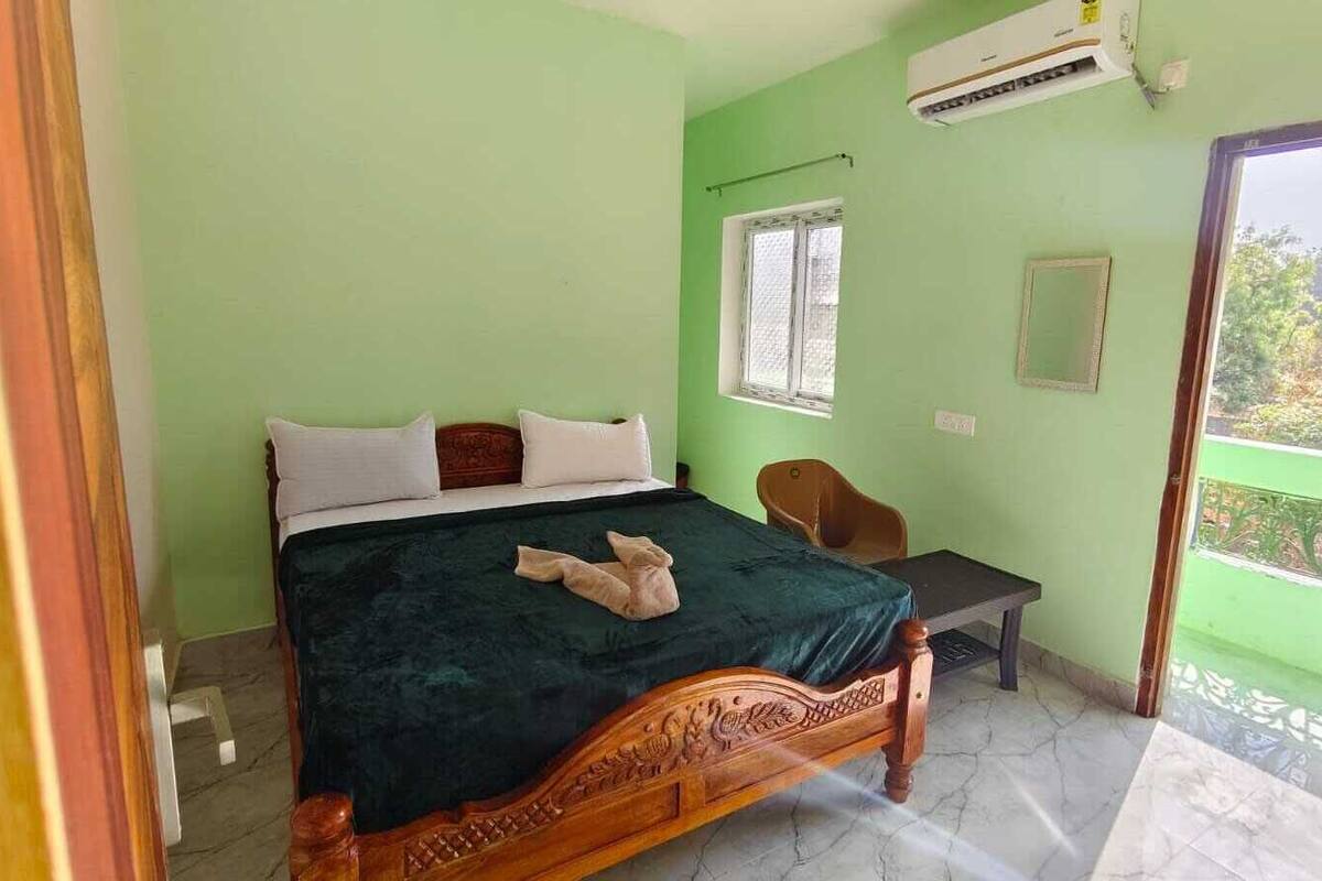 A bedroom with a bed, a chair, a small table, a window, and an air conditioning unit.