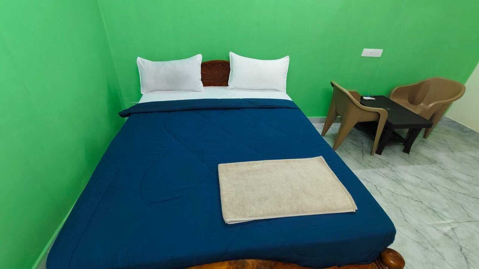 Una cama individual con colcha azul y almohadas blancas, una pequeña silla de madera y una mesita con una silla.