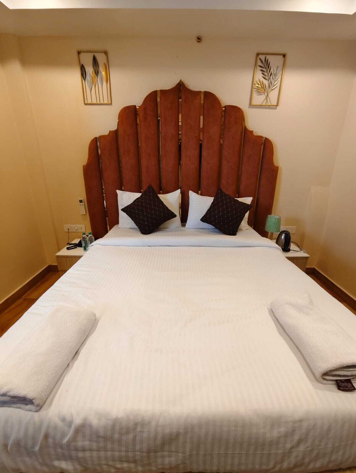 A hotel room with a large bed, a wooden headboard, two wall-mounted paintings, a bedside table with a lamp, and a folded towel on the bed.