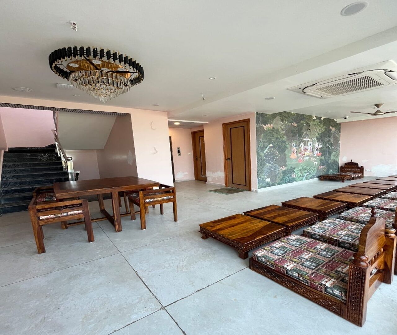 A spacious room with a dining table and chairs, a long sofa with patterned cushions, a chandelier, and a staircase leading to another level.