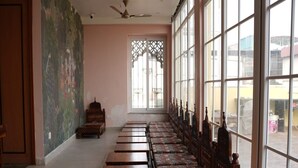 A room with a long row of wooden benches and a patterned sofa, large windows, and a ceiling fan. - Atithi Sevalay (Varanasi)