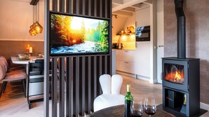 A modern living room with a TV mounted on a wooden panel, a fireplace, and a dining area with a table and chairs. - Casa Vida Verde - Luxury cottage by water & nature (Gramsbergen)