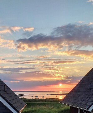 Ein Sonnenuntergangsblick von einem Gebäude mit geneigtem Dach, einem grasbewachsen Vordergrund und einem Gewässer in der Ferne.