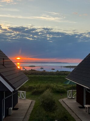 Ein Sonnenuntergang, der von einer Holzhütte mit einer Holzterrasse und einem grasbewachsen Vordergrund aus gesehen wird.