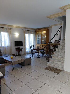 A living room with a sofa, a coffee table, a dining table with chairs, a staircase, and a TV mounted on the wall. - House T4 center Bastia - terrace, comfort and ideal location (BASTIA)