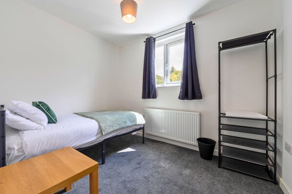 A compact room with a single bed, a small table, a radiator, a window with curtains, and a metal shelf unit.