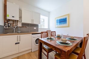 A compact kitchen with a built-in washer, a dining table set for four, and a framed artwork on the wall. - 6 Bedroom House for up to 10 People (Merthyr Tydfil)