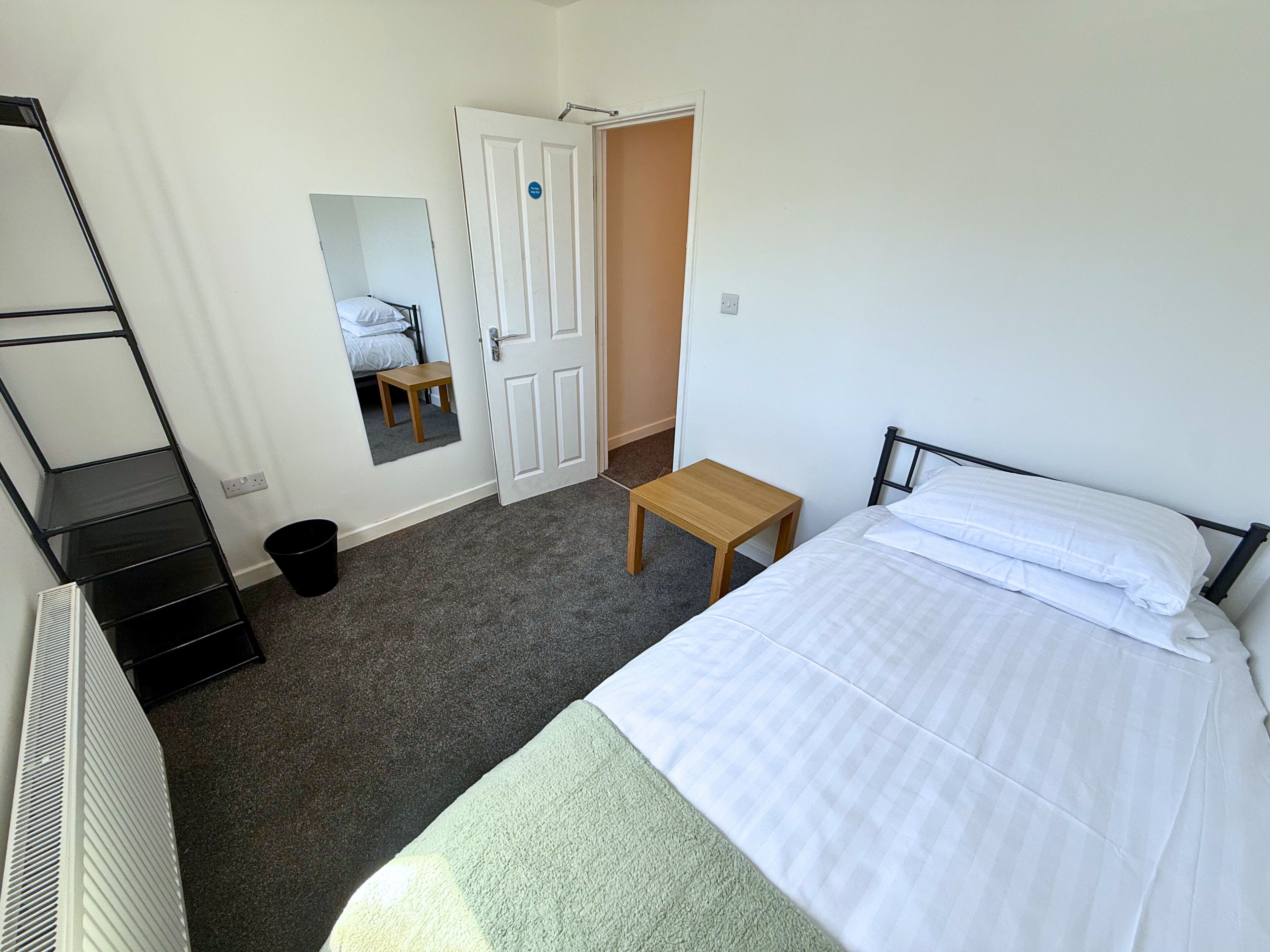 A bedroom with a bed, a small table, a mirror, a radiator, and a door leading to another room.