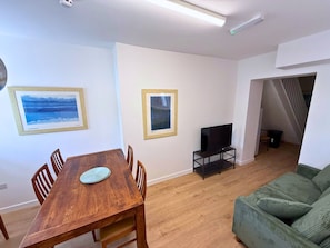 A living room with a wooden dining table, two framed artworks, a television on a stand, and a green sofa. - 6 Bedroom House for up to 10 People (Merthyr Tydfil)