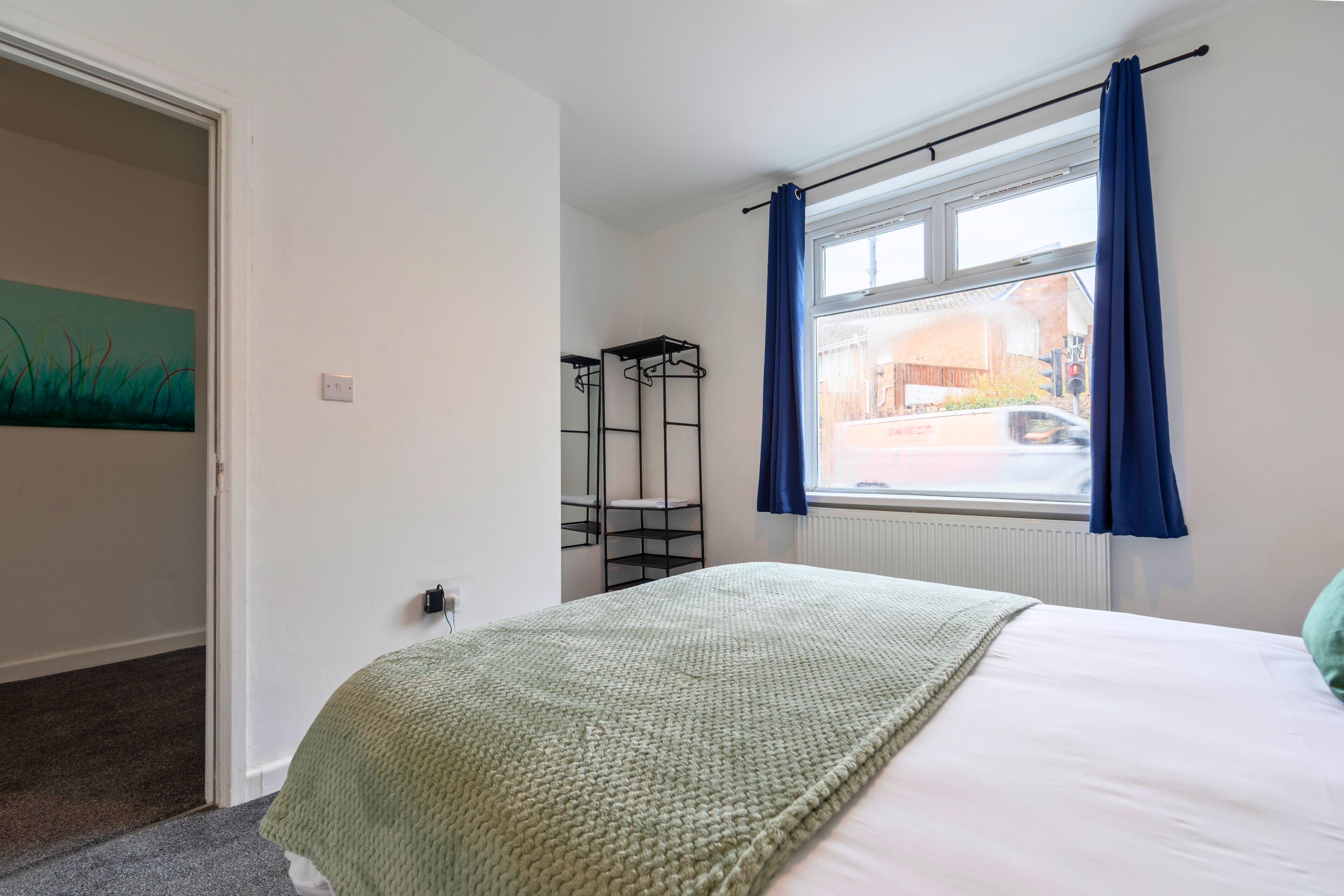 A bedroom with a bed, a window with curtains, a radiator, and a wall-mounted shelf.