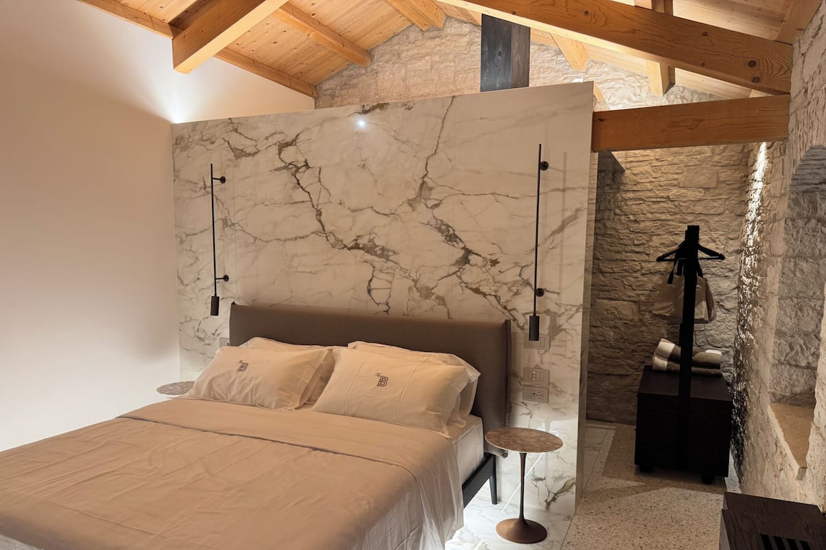 Un dormitorio con cama, pared de mármol, vigas de madera, pared de piedra y una pequeña mesa.