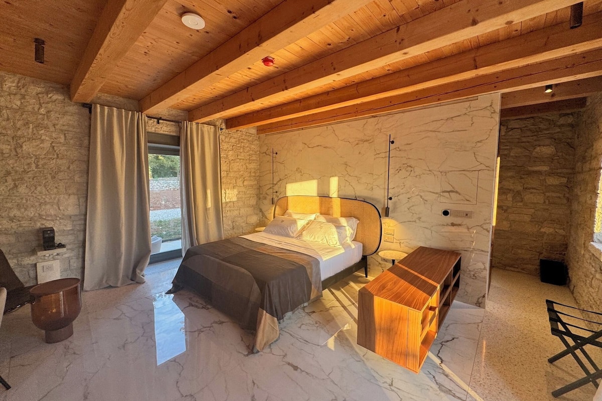 Un dormitorio con cama, mesita de noche, lámpara, paredes de piedra, vigas de madera en el techo y vistas por una ventana.