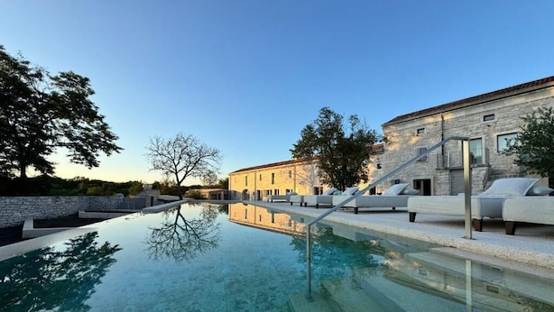Une piscine avec des marches menant à un bâtiment doté d’une façade en pierre et d’un espace extérieur aménagé pour s’asseoir.