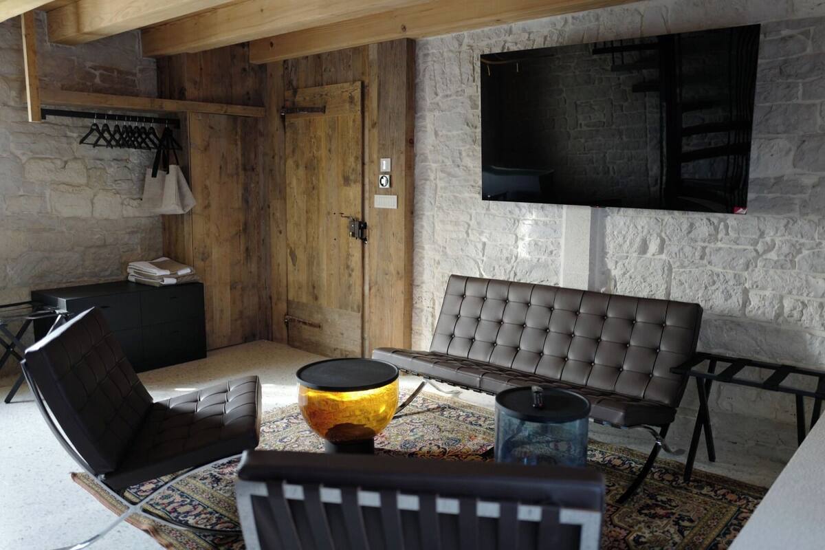 Una habitación con pared de piedra, puerta de madera, televisor de pantalla plana, sofá, sillón, mesita y lámpara.