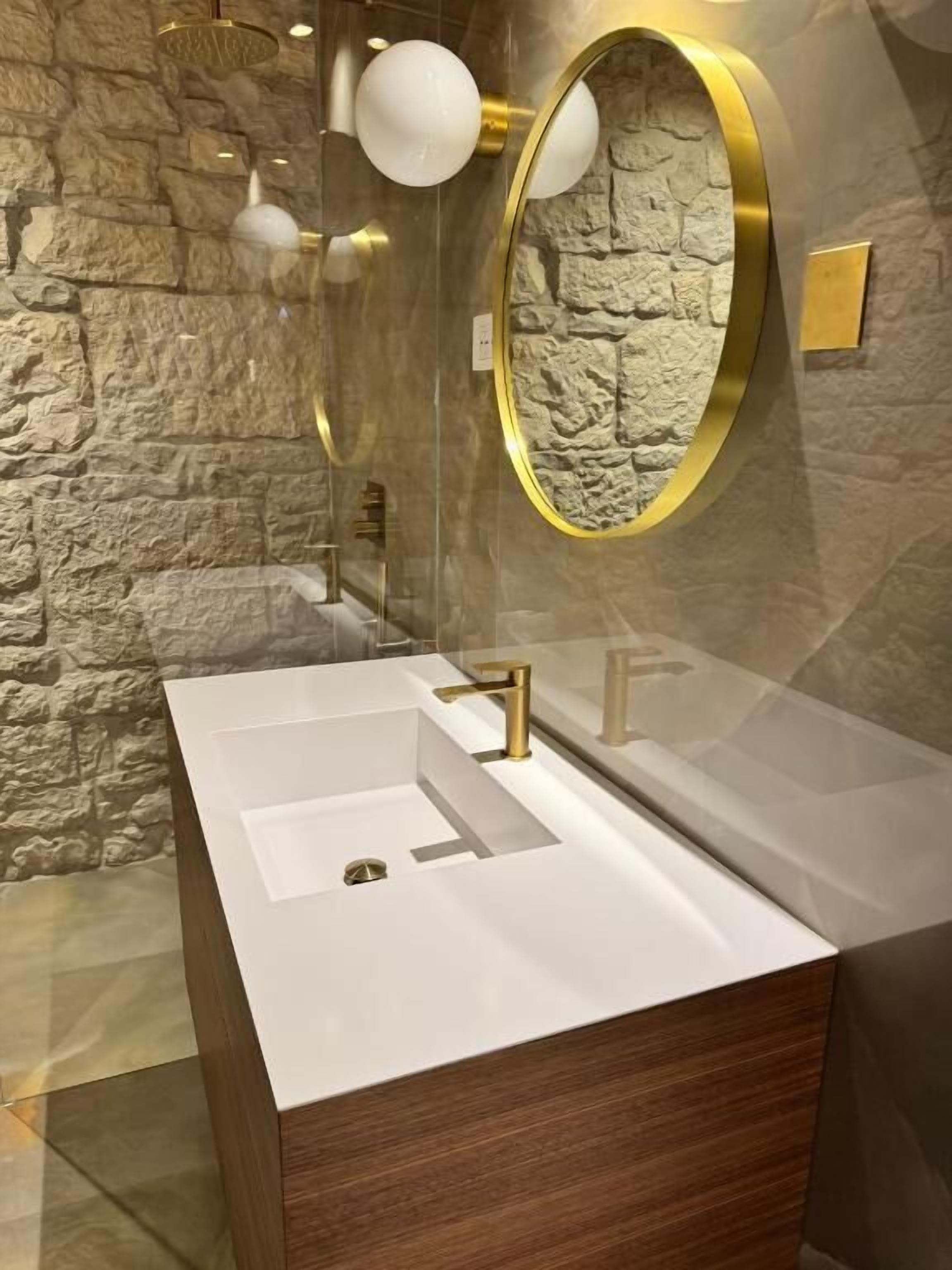 A bathroom with a white sink, gold faucets, a round mirror with a gold frame, and a stone-textured wall.