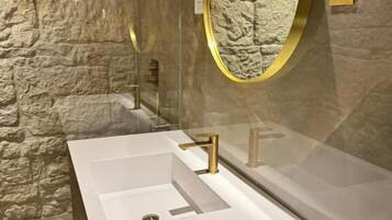 A bathroom with a white sink, gold faucets, a round mirror with a gold frame, and a stone-textured wall.