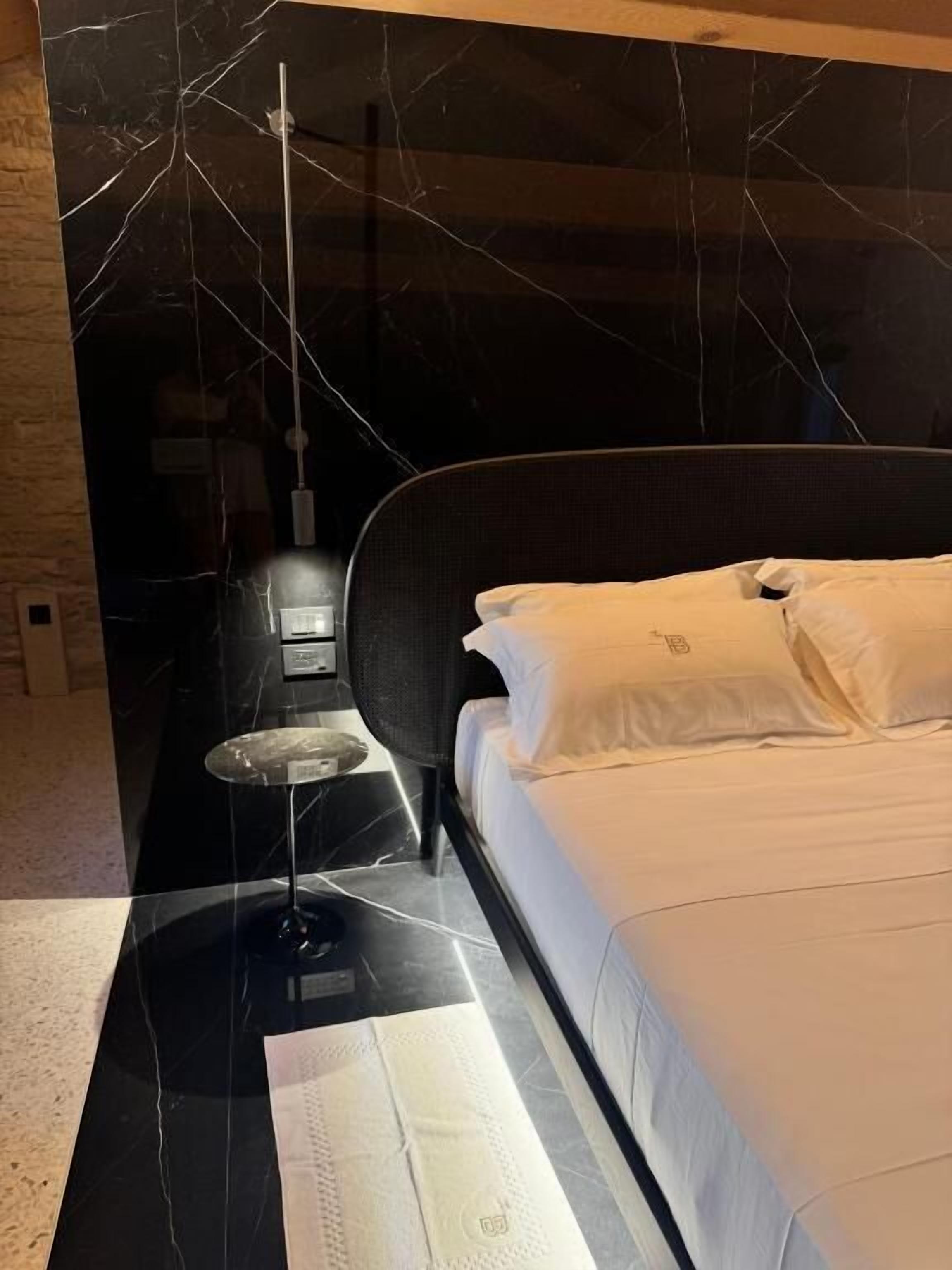 A modern bedroom with a black marble wall, a bed with white bedding, a small bedside table, and a hanging light fixture.