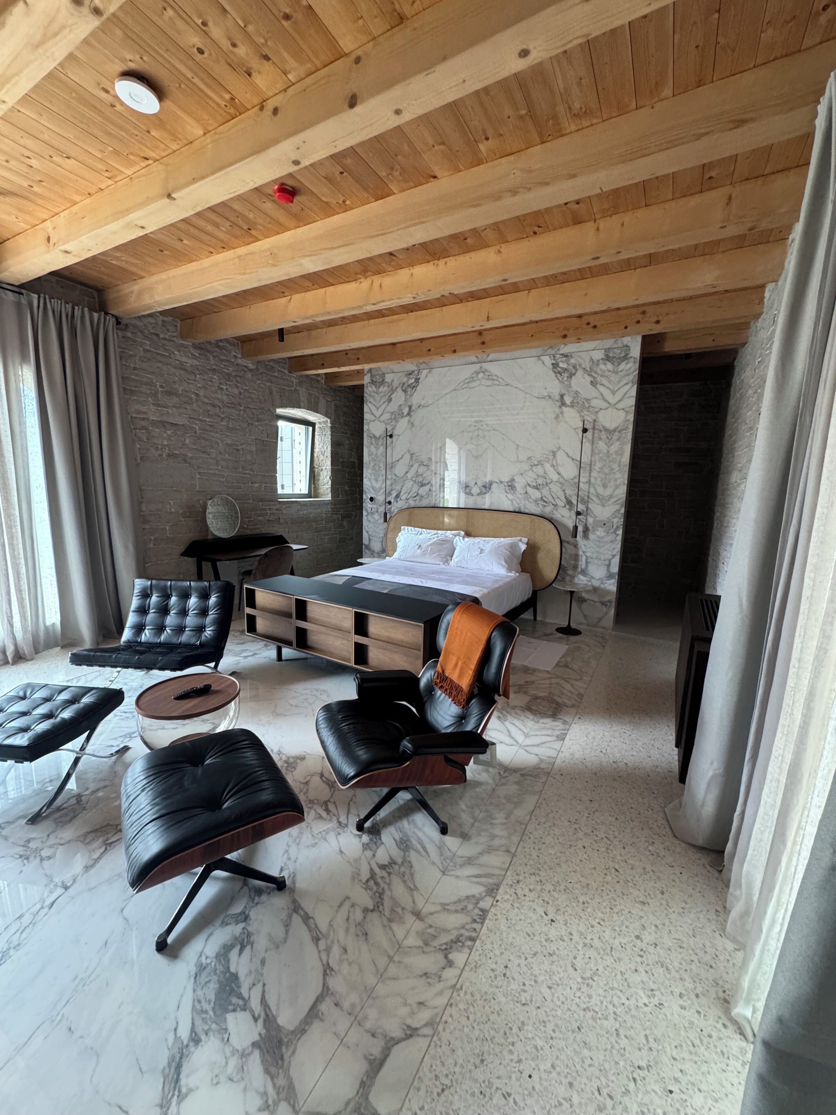 Une chambre à coucher moderne avec un lit, deux fauteuils en cuir, une petite table et un mur en pierre.