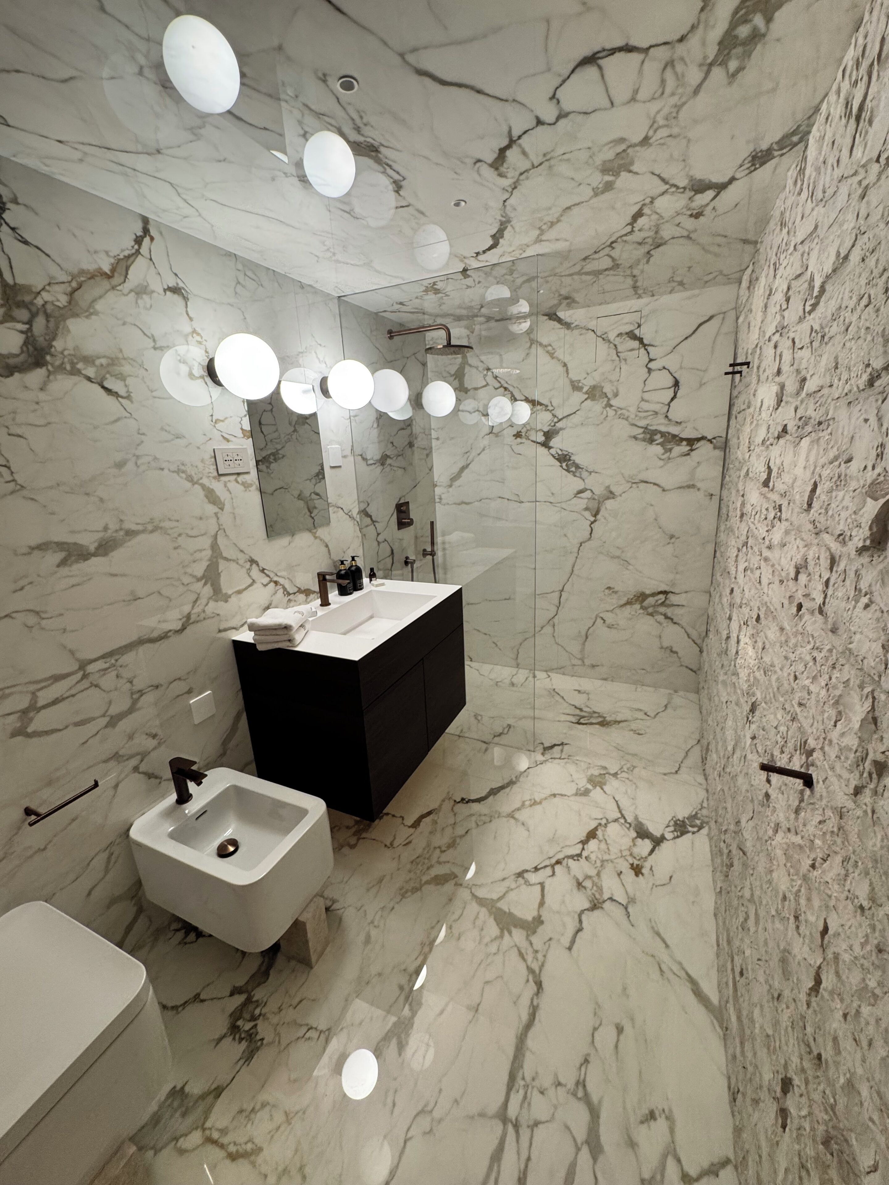 A bathroom with a marble floor, a white sink, a black vanity, and a mirror with lights above.