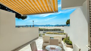 Un espace extérieur aménagé, abrité et doté de mobilier en osier et d’une table, offrant une vue sur une plage parsemée de parasols et fréquentée par des personnes.