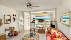 Un salon avec une table basse en verre, un canapé à rayures, un ventilateur de plafond et une vue sur l’extérieur, visible à travers une porte vitrée.