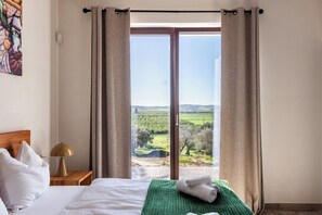 A hotel room with a bed, a nightstand, a lamp, and a view of a green landscape through large windows. - Villa Maddalena - Three Bedroom Villa, Sleeps 6 (Noto)