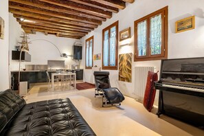 A living room with a black leather sofa, a piano, a dining area with a table and chairs, a kitchen in the background, and a window with curtains. - Cà Leon, Charming Loft in the Heart of the City (Venezia)