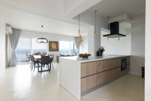 A modern kitchen with a large island, dining area with chairs, and a view of the sea. - Elegant seafront 2BR apartment, stunning sea views, just steps to shore! (Oropioi)