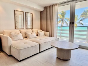 Ein modernes Wohnzimmer mit einem beigefarbenen Ecksofa, einem runden Couchtisch und einer großen Schiebeglasdoor, die auf einen Balkon mit Blick aufs Meer und Palmen führt.