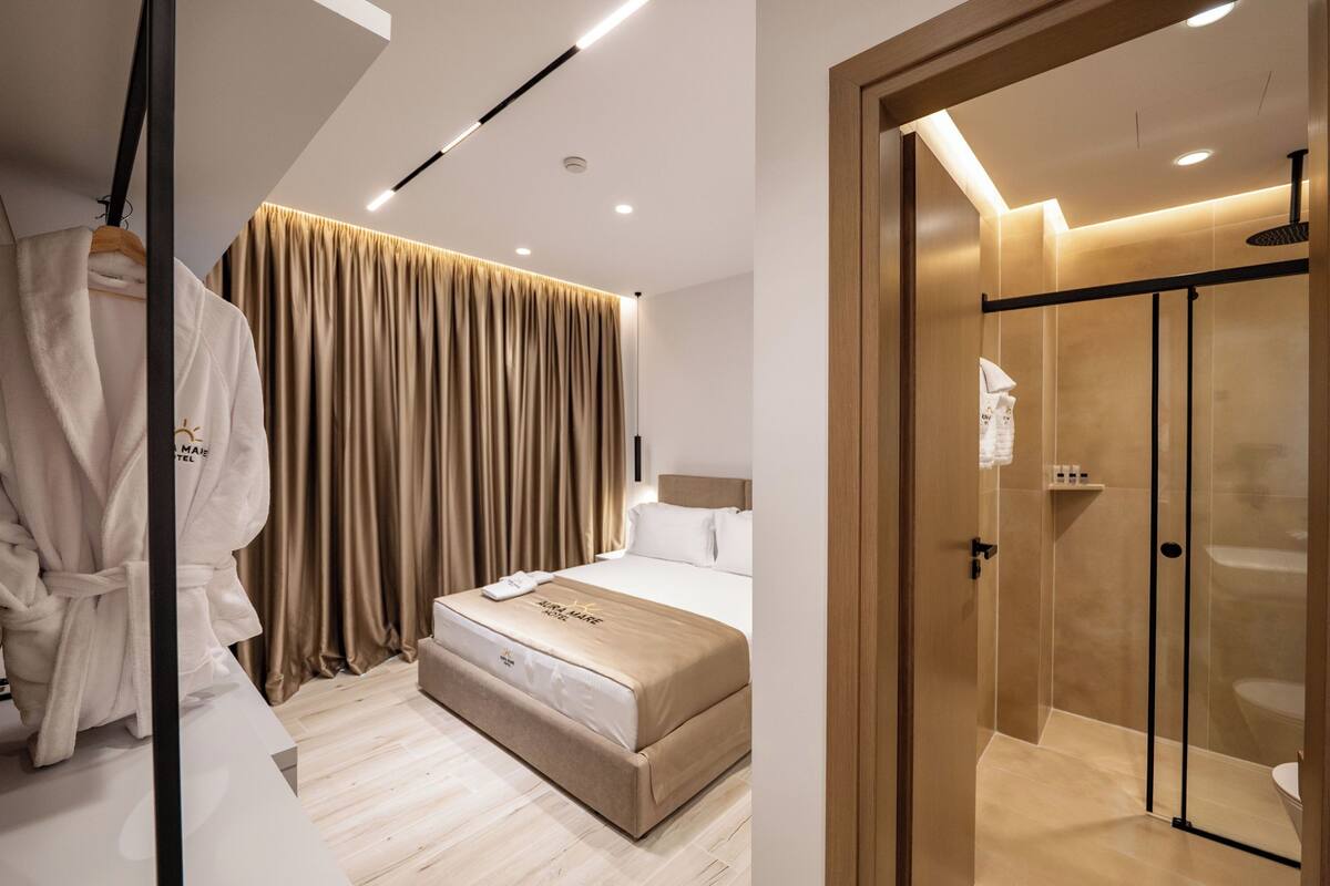 Una habitación de hotel moderna con cama, perchero para bata, ventana grande con cortinas y cabina de ducha con paredes de vidrio.