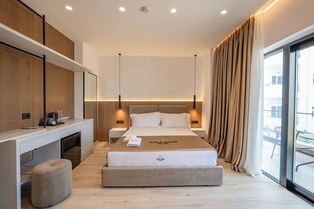 Una habitación de hotel moderna con cama, mesitas de noche, escritorio y un amplio ventanal con cortinas.