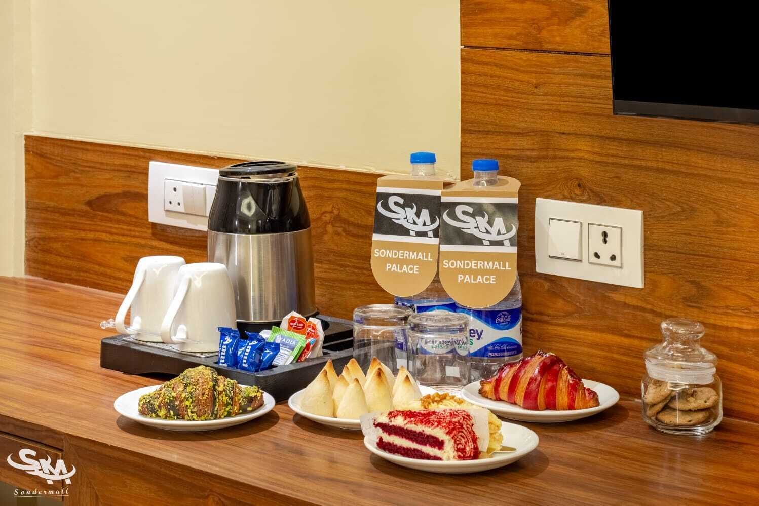 A hotel reception area with a coffee maker, water dispensers, and a variety of food items on the counter.