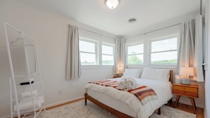 Une chambre à coucher avec un lit, une table de chevet, une lampe, une fenêtre, un mur blanc et un plancher en bois.