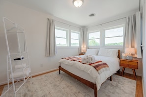 Ein Schlafzimmer mit Bett, Nachttisch, Lampe, Fenster, weißer Wand und Holzboden.